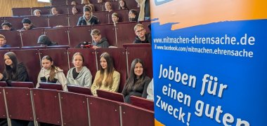 Ein Banner des Projekt "Mitmachen ist Ehrensache" ist im Vordergrund. Dahinter sitzen Schüler und warten auf den Beginn des Vortrags.