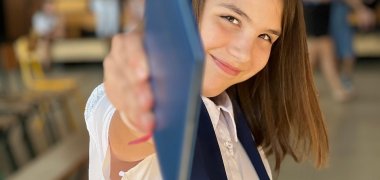 Eine junge Frau zeigt mit einem blaue dünnen Buch in die Kamera
