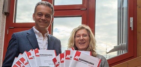 Bürgermeister links und Öffentlichkeitsarbeit rechts präsentieren den Jahresbericht. Sie lachen in die Kamera.