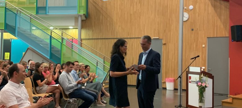 Bürgermeister Schellinger übereicht Frau Wiedmann einen Gutschein. Im Hintergrund das applaudierende Publikum.