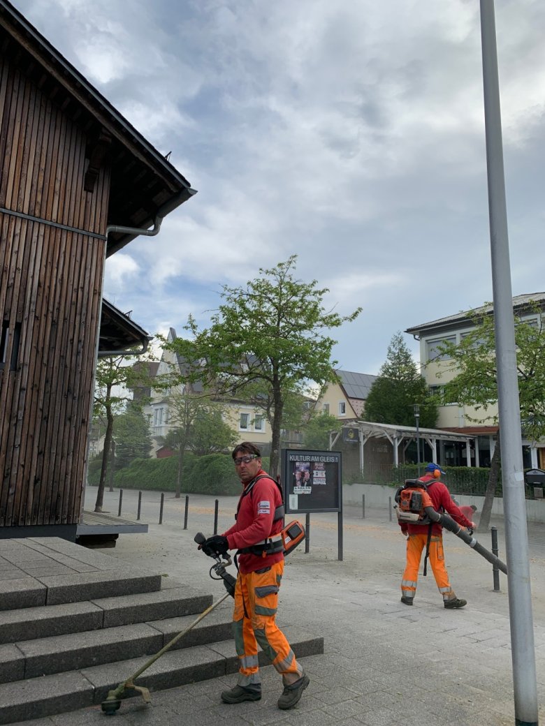 Bahnhofsvorplatz Meckenbeuren mit Blumen und Menschen, die saubermachen